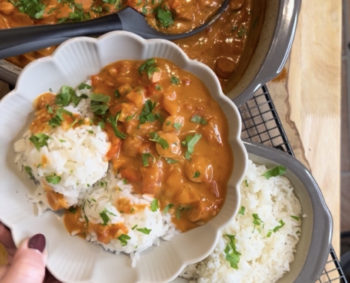 Ein fertig angerichtetes Würstchengulasch mit fluffigem Reis in einer weißen, blütenförmigen Schale. Im Hintergrund steht das Gulasch im grauen Ofenmeister und der Reis im kleinen Zaubermeister von Pampered Chef auf einem Kuchengitter. Die Szene ist mit frischer Petersilie, Kirschtomaten und einer roten Paprika auf einem rustikalen Holzbrett dekoriert.