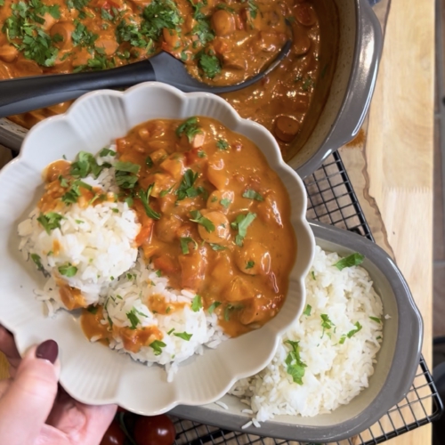 Ein fertig angerichtetes Würstchengulasch mit fluffigem Reis in einer weißen, blütenförmigen Schale. Im Hintergrund steht das Gulasch im grauen Ofenmeister und der Reis im kleinen Zaubermeister von Pampered Chef auf einem Kuchengitter. Die Szene ist mit frischer Petersilie, Kirschtomaten und einer roten Paprika auf einem rustikalen Holzbrett dekoriert.