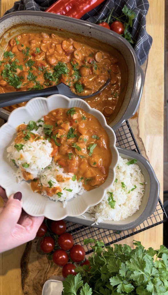 Ein fertig angerichtetes Würstchengulasch mit fluffigem Reis in einer weißen, blütenförmigen Schale. Im Hintergrund steht das Gulasch im grauen Ofenmeister und der Reis im kleinen Zaubermeister von Pampered Chef auf einem Kuchengitter. Die Szene ist mit frischer Petersilie, Kirschtomaten und einer roten Paprika auf einem rustikalen Holzbrett dekoriert.