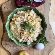 Hausgemachter Kartoffelsalat mit Äpfeln, Fleischwurst und Mayonnaise in einer grünen, blattförmigen Schüssel auf einem Holzbrett, dekoriert mit frischen Äpfeln und Gewürzgurken.