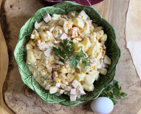 Hausgemachter Kartoffelsalat mit Äpfeln, Fleischwurst und Mayonnaise in einer grünen, blattförmigen Schüssel auf einem Holzbrett, dekoriert mit frischen Äpfeln und Gewürzgurken.